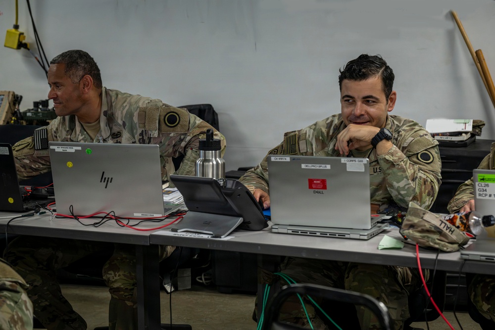 Two U.S. Army soldiers in OCP (Operational Camouflage Pattern) uniforms are seated at a table working on laptops in what appears to be a tactical operations center or field command post. The soldier on the right is smiling and appears relaxed, while the soldier on the left is focused on his HP laptop screen. Multiple computers, cables, and military equipment are visible on the workspace.