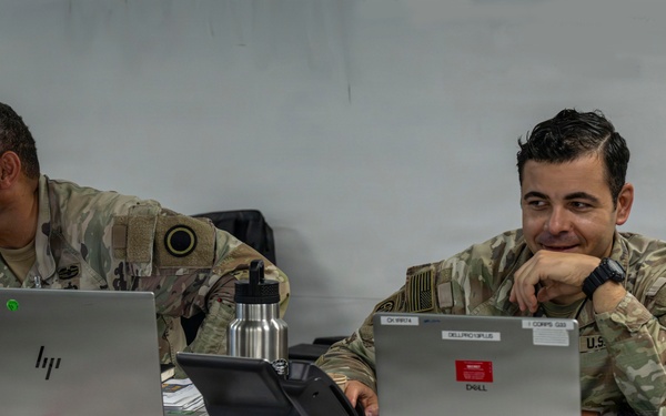 Two U.S. Army soldiers in OCP (Operational Camouflage Pattern) uniforms are seated at a table working on laptops in what appears to be a tactical operations center or field command post. The soldier on the right is smiling and appears relaxed, while the soldier on the left is focused on his HP laptop screen. Multiple computers, cables, and military equipment are visible on the workspace.