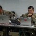 Two U.S. Army soldiers in OCP (Operational Camouflage Pattern) uniforms are seated at a table working on laptops in what appears to be a tactical operations center or field command post. The soldier on the right is smiling and appears relaxed, while the soldier on the left is focused on his HP laptop screen. Multiple computers, cables, and military equipment are visible on the workspace.
