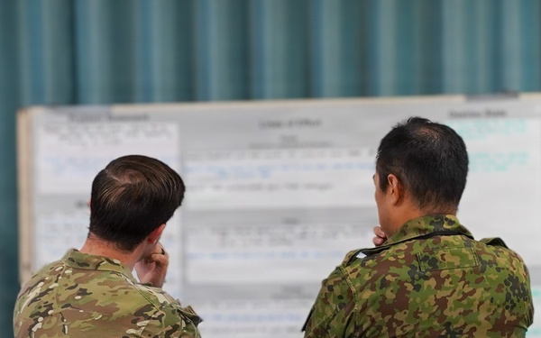 Two military personnel in different camouflage uniforms are standing with their backs to the camera, studying a large whiteboard or briefing board covered in text and charts. A third individual is partially visible in the lower foreground. The setting appears to be an indoor briefing or planning room with teal curtains in the background.