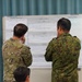 Two military personnel in different camouflage uniforms are standing with their backs to the camera, studying a large whiteboard or briefing board covered in text and charts. A third individual is partially visible in the lower foreground. The setting appears to be an indoor briefing or planning room with teal curtains in the background.