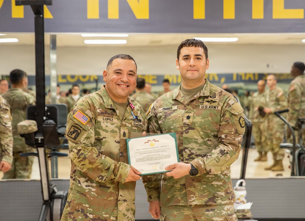 Two U.S. Army soldiers pose together holding a certificate in what appears to be a military fitness facility. The soldier on the left, identified by his name tape as 'LOPEZ,' is wearing a medal and holds the certificate alongside the soldier on the right, identified as 'ZIMMER,' who bears a Lieutenant Colonel oak leaf insignia. The certificate appears to be a Meritorious Service Medal award citation.