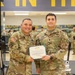 Two U.S. Army soldiers pose together holding a certificate in what appears to be a military fitness facility. The soldier on the left, identified by his name tape as 'LOPEZ,' is wearing a medal and holds the certificate alongside the soldier on the right, identified as 'ZIMMER,' who bears a Lieutenant Colonel oak leaf insignia. The certificate appears to be a Meritorious Service Medal award citation.