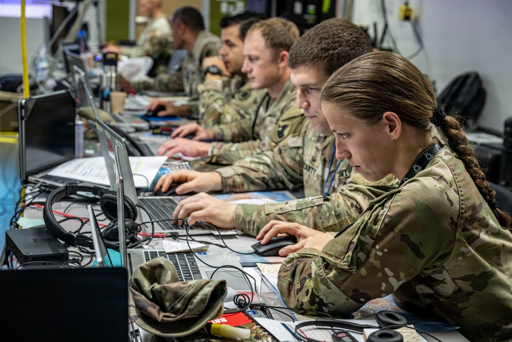 Military personnel in MultiCam/OCP camouflage uniforms are working at a long desk equipped with multiple laptops and computer equipment in what appears to be a tactical operations center or command post. At least four soldiers are visible, focused on their screens in a row, suggesting a coordinated cyber, intelligence, or command-and-control operation. The environment is densely equipped with cables, headsets, documents, and various electronic devices.