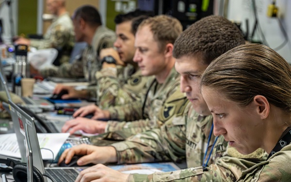 Military personnel in MultiCam/OCP camouflage uniforms are working at a long desk equipped with multiple laptops and computer equipment in what appears to be a tactical operations center or command post. At least four soldiers are visible, focused on their screens in a row, suggesting a coordinated cyber, intelligence, or command-and-control operation. The environment is densely equipped with cables, headsets, documents, and various electronic devices.