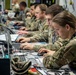 Military personnel in MultiCam/OCP camouflage uniforms are working at a long desk equipped with multiple laptops and computer equipment in what appears to be a tactical operations center or command post. At least four soldiers are visible, focused on their screens in a row, suggesting a coordinated cyber, intelligence, or command-and-control operation. The environment is densely equipped with cables, headsets, documents, and various electronic devices.