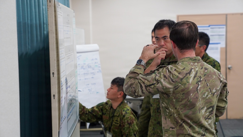 Military personnel in two distinct camouflage uniforms are gathered in what appears to be a briefing or planning room, studying documents and charts posted on a board. The scene suggests a joint military exercise or planning session involving soldiers from at least two different nations. Personnel are engaged in focused discussion and review of tactical or operational materials.