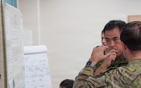 Military personnel in two distinct camouflage uniforms are gathered in what appears to be a briefing or planning room, studying documents and charts posted on a board. The scene suggests a joint military exercise or planning session involving soldiers from at least two different nations. Personnel are engaged in focused discussion and review of tactical or operational materials.