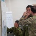 Military personnel in two distinct camouflage uniforms are gathered in what appears to be a briefing or planning room, studying documents and charts posted on a board. The scene suggests a joint military exercise or planning session involving soldiers from at least two different nations. Personnel are engaged in focused discussion and review of tactical or operational materials.