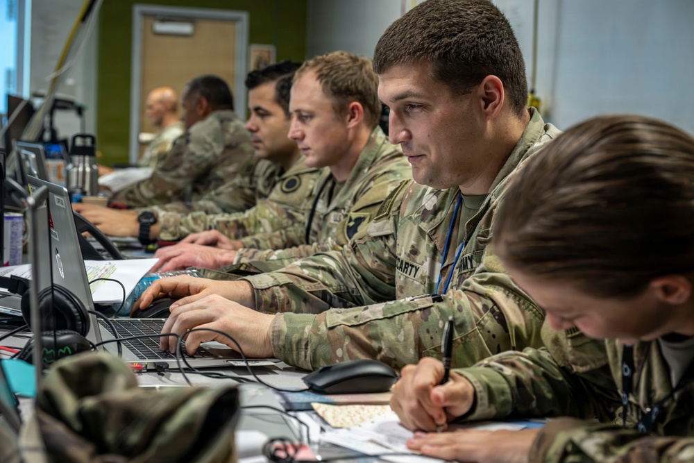A group of U.S. Army soldiers in OCP (Operational Camouflage Pattern) uniforms are working at a row of laptops in what appears to be a military operations or command center. The soldiers are focused on their screens and paperwork, suggesting a training exercise, planning session, or operational briefing. The environment is an indoor facility with multiple workstations set up in a linear arrangement.