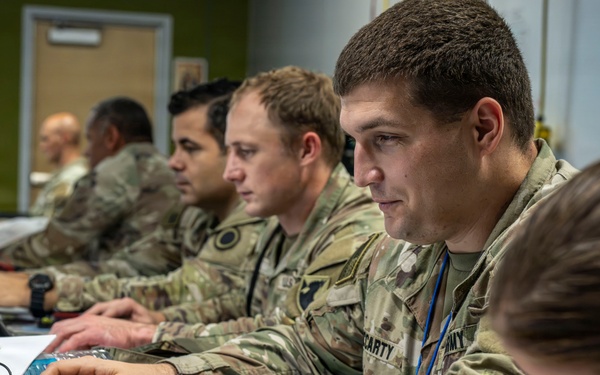 A group of U.S. Army soldiers in OCP (Operational Camouflage Pattern) uniforms are working at a row of laptops in what appears to be a military operations or command center. The soldiers are focused on their screens and paperwork, suggesting a training exercise, planning session, or operational briefing. The environment is an indoor facility with multiple workstations set up in a linear arrangement.