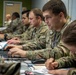 A group of U.S. Army soldiers in OCP (Operational Camouflage Pattern) uniforms are working at a row of laptops in what appears to be a military operations or command center. The soldiers are focused on their screens and paperwork, suggesting a training exercise, planning session, or operational briefing. The environment is an indoor facility with multiple workstations set up in a linear arrangement.