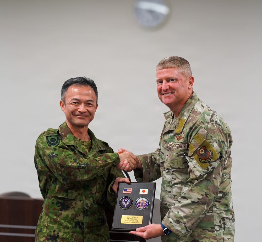 Two military officers, one from the Japan Ground Self-Defense Force and one from the U.S. Air Force, are shaking hands while jointly holding a commemorative plaque. The plaque displays both the American and Japanese flags along with two military challenge coins, and bears text reading 'U.S.-Japan Bilateral Engagement.' This appears to be a formal bilateral military engagement ceremony or exchange event.
