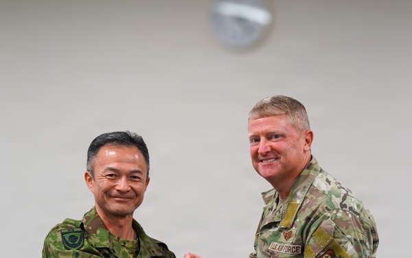 Two military officers, one from the Japan Ground Self-Defense Force and one from the U.S. Air Force, are shaking hands while jointly holding a commemorative plaque. The plaque displays both the American and Japanese flags along with two military challenge coins, and bears text reading 'U.S.-Japan Bilateral Engagement.' This appears to be a formal bilateral military engagement ceremony or exchange event.