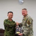 Two military officers, one from the Japan Ground Self-Defense Force and one from the U.S. Air Force, are shaking hands while jointly holding a commemorative plaque. The plaque displays both the American and Japanese flags along with two military challenge coins, and bears text reading 'U.S.-Japan Bilateral Engagement.' This appears to be a formal bilateral military engagement ceremony or exchange event.