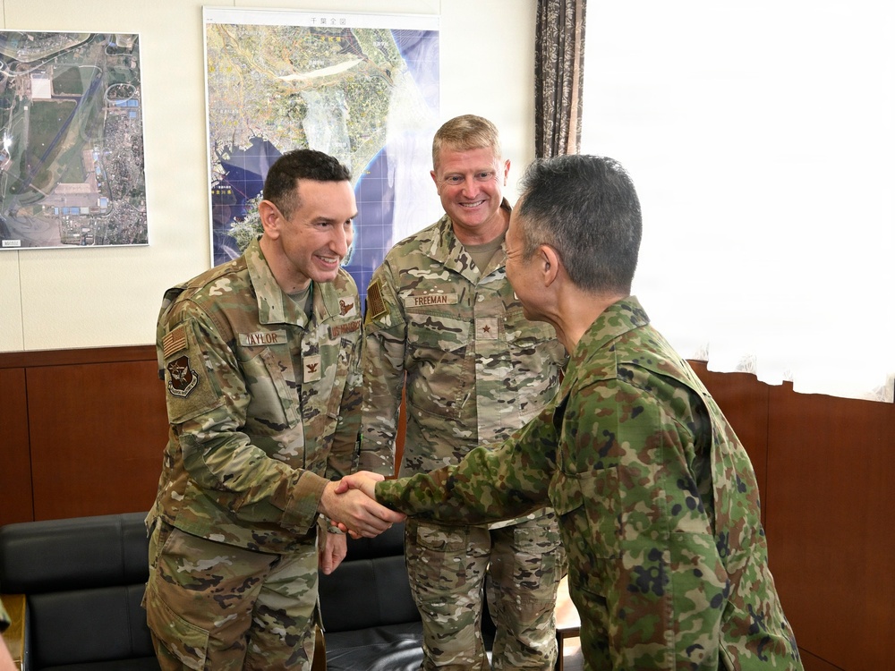 Three military personnel are engaged in a greeting exchange in what appears to be an office or briefing room. Two individuals wearing US military OCP (Operational Camouflage Pattern) uniforms with visible name tapes 'TAYLOR' and 'FREEMAN' are meeting with a third individual in a different camouflage pattern consistent with Japan Ground Self-Defense Force (JGSDF) uniform. A handshake is taking place between TAYLOR and the JGSDF member, suggesting a formal bilateral military meeting.