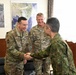 Three military personnel are engaged in a greeting exchange in what appears to be an office or briefing room. Two individuals wearing US military OCP (Operational Camouflage Pattern) uniforms with visible name tapes 'TAYLOR' and 'FREEMAN' are meeting with a third individual in a different camouflage pattern consistent with Japan Ground Self-Defense Force (JGSDF) uniform. A handshake is taking place between TAYLOR and the JGSDF member, suggesting a formal bilateral military meeting.