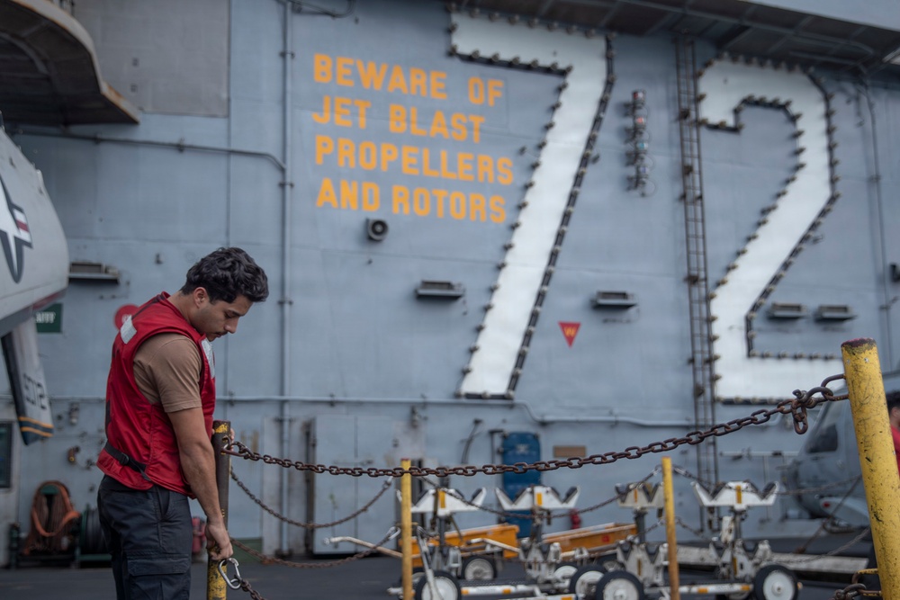 A sailor wearing a red safety vest works on the flight deck of USS Abraham Lincoln (CVN-72), a U.S. Navy aircraft carrier. He is handling a heavy chain, likely securing or unsecuring aircraft tie-downs. The ship's island superstructure displays the hull number '72' and a yellow safety warning reading 'BEWARE OF JET BLAST PROPELLERS AND ROTORS.'