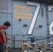 A sailor wearing a red safety vest works on the flight deck of USS Abraham Lincoln (CVN-72), a U.S. Navy aircraft carrier. He is handling a heavy chain, likely securing or unsecuring aircraft tie-downs. The ship's island superstructure displays the hull number '72' and a yellow safety warning reading 'BEWARE OF JET BLAST PROPELLERS AND ROTORS.'