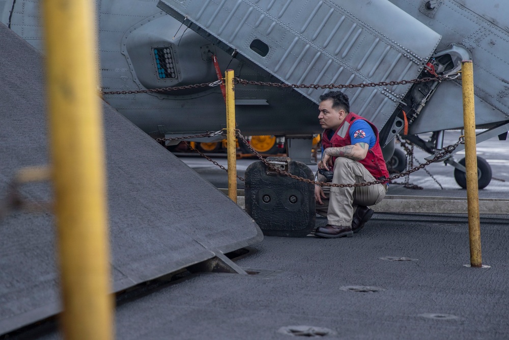 A male technician or deck crew member wearing a red vest over a blue shirt and khaki pants is crouching on what appears to be a naval aircraft carrier flight deck. He is positioned near a chained-down military aircraft, likely performing an inspection or maintenance check. Yellow safety posts and heavy chains are used to secure the aircraft to the deck.