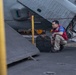 A male technician or deck crew member wearing a red vest over a blue shirt and khaki pants is crouching on what appears to be a naval aircraft carrier flight deck. He is positioned near a chained-down military aircraft, likely performing an inspection or maintenance check. Yellow safety posts and heavy chains are used to secure the aircraft to the deck.