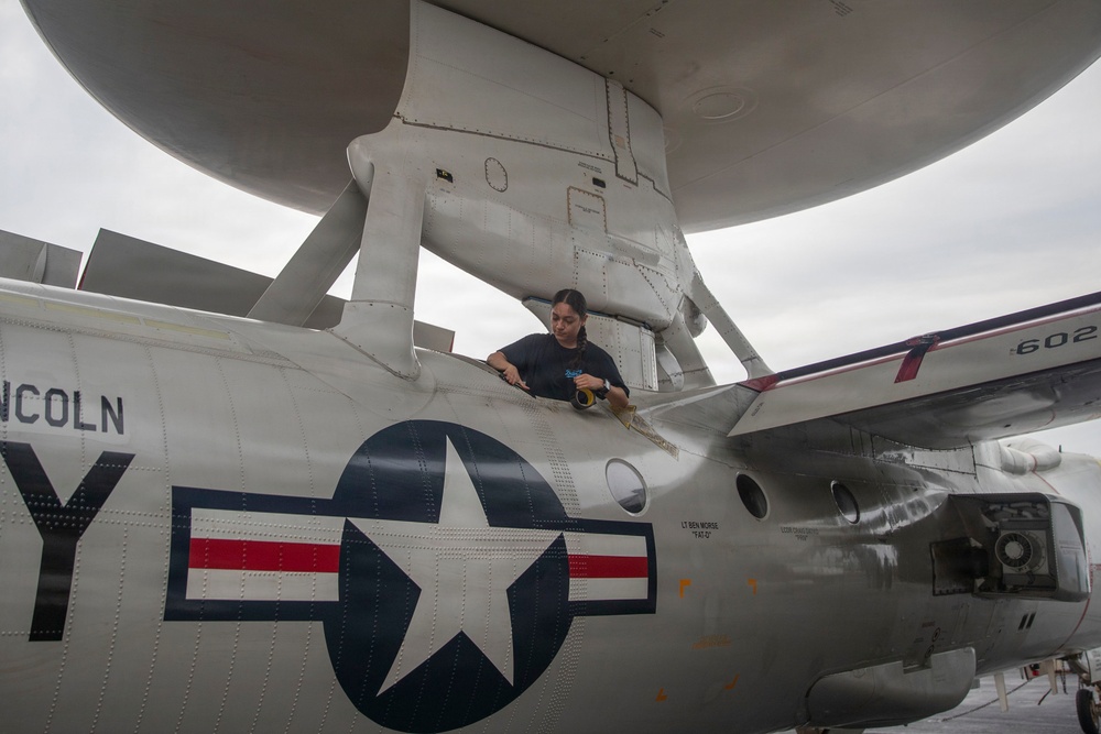 A technician or maintenance worker is performing work on top of a U.S. Navy aircraft, identifiable by the distinctive rotodome (radar disc) mounted above the fuselage, characteristic of an E-2 Hawkeye airborne early warning aircraft. The aircraft bears U.S. military insignia including the standard roundel (star and bar) and partial text reading 'LINCOLN' and tail number '602'. The worker appears to be applying tape or performing surface maintenance on the aircraft's upper fuselage.