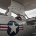 A technician or maintenance worker is performing work on top of a U.S. Navy aircraft, identifiable by the distinctive rotodome (radar disc) mounted above the fuselage, characteristic of an E-2 Hawkeye airborne early warning aircraft. The aircraft bears U.S. military insignia including the standard roundel (star and bar) and partial text reading 'LINCOLN' and tail number '602'. The worker appears to be applying tape or performing surface maintenance on the aircraft's upper fuselage.
