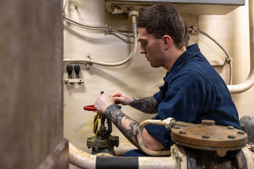 A young male technician wearing a navy blue uniform is working on industrial piping or valve equipment in what appears to be a mechanical room or engine room. He is focused on manipulating a red-handled valve with yellow safety tags attached. The environment features aged, cream-colored walls with conduit wiring and heavy-duty flanged pipe fittings.