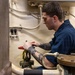 A young male technician wearing a navy blue uniform is working on industrial piping or valve equipment in what appears to be a mechanical room or engine room. He is focused on manipulating a red-handled valve with yellow safety tags attached. The environment features aged, cream-colored walls with conduit wiring and heavy-duty flanged pipe fittings.