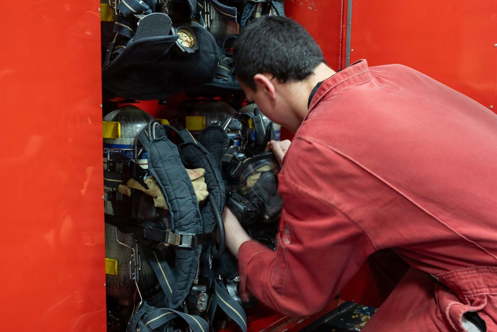 A young person wearing a red uniform or jumpsuit is leaning into a red storage compartment, handling firefighting equipment. The compartment contains multiple Self-Contained Breathing Apparatus (SCBA) units with harnesses, masks, and pressure gauges. This appears to be a fire truck equipment bay or fire station gear storage area.