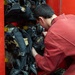 A young person wearing a red uniform or jumpsuit is leaning into a red storage compartment, handling firefighting equipment. The compartment contains multiple Self-Contained Breathing Apparatus (SCBA) units with harnesses, masks, and pressure gauges. This appears to be a fire truck equipment bay or fire station gear storage area.