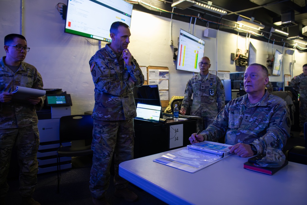 Military personnel in U.S. Army/Air Force camouflage uniforms are gathered in what appears to be a tactical operations center (TOC) or command post. Multiple screens displaying data dashboards are visible on the walls, and personnel appear to be engaged in a briefing or planning session. The environment is dimly lit with overhead fluorescent lighting and features multiple workstations.
