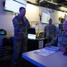 Military personnel in U.S. Army/Air Force camouflage uniforms are gathered in what appears to be a tactical operations center (TOC) or command post. Multiple screens displaying data dashboards are visible on the walls, and personnel appear to be engaged in a briefing or planning session. The environment is dimly lit with overhead fluorescent lighting and features multiple workstations.