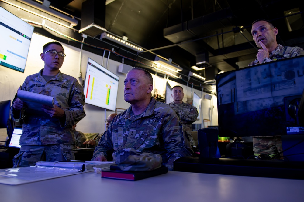 Several U.S. Army personnel are gathered in what appears to be a military operations or command center, reviewing data displayed on large wall-mounted screens. The central figure, identified by name tape as 'LOGAN,' is seated at a workstation with a computer monitor, while colleagues stand nearby holding documents and observing the displays. The environment suggests a briefing or situational awareness review session.