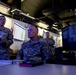 Several U.S. Army personnel are gathered in what appears to be a military operations or command center, reviewing data displayed on large wall-mounted screens. The central figure, identified by name tape as 'LOGAN,' is seated at a workstation with a computer monitor, while colleagues stand nearby holding documents and observing the displays. The environment suggests a briefing or situational awareness review session.
