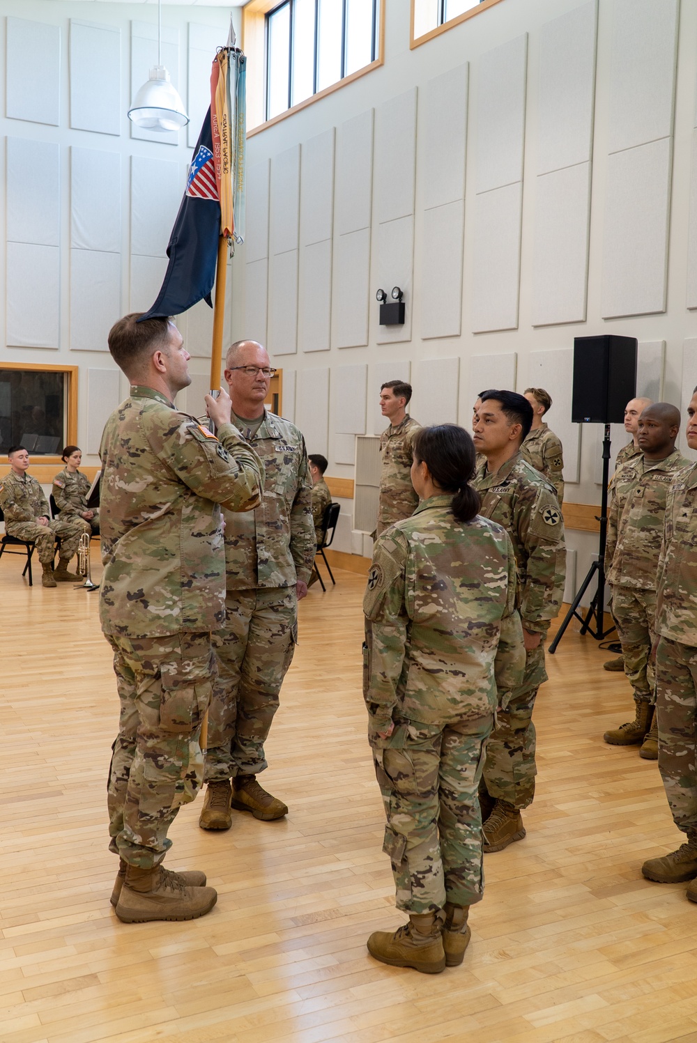 A U.S. Army military ceremony is taking place indoors in what appears to be a multipurpose hall with hardwood floors and acoustic wall panels. Two soldiers in the foreground are engaged in a guidon or colors transfer, with one soldier holding a unit flag on a staff while facing another. Additional soldiers stand in formation observing the ceremony, with seated personnel visible in the background.