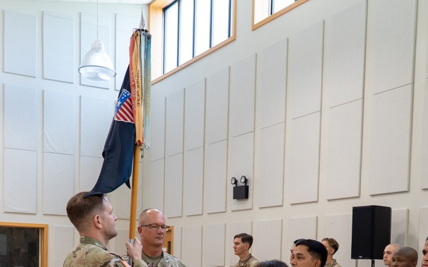 A U.S. Army military ceremony is taking place indoors in what appears to be a multipurpose hall with hardwood floors and acoustic wall panels. Two soldiers in the foreground are engaged in a guidon or colors transfer, with one soldier holding a unit flag on a staff while facing another. Additional soldiers stand in formation observing the ceremony, with seated personnel visible in the background.
