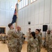 A U.S. Army military ceremony is taking place indoors in what appears to be a multipurpose hall with hardwood floors and acoustic wall panels. Two soldiers in the foreground are engaged in a guidon or colors transfer, with one soldier holding a unit flag on a staff while facing another. Additional soldiers stand in formation observing the ceremony, with seated personnel visible in the background.