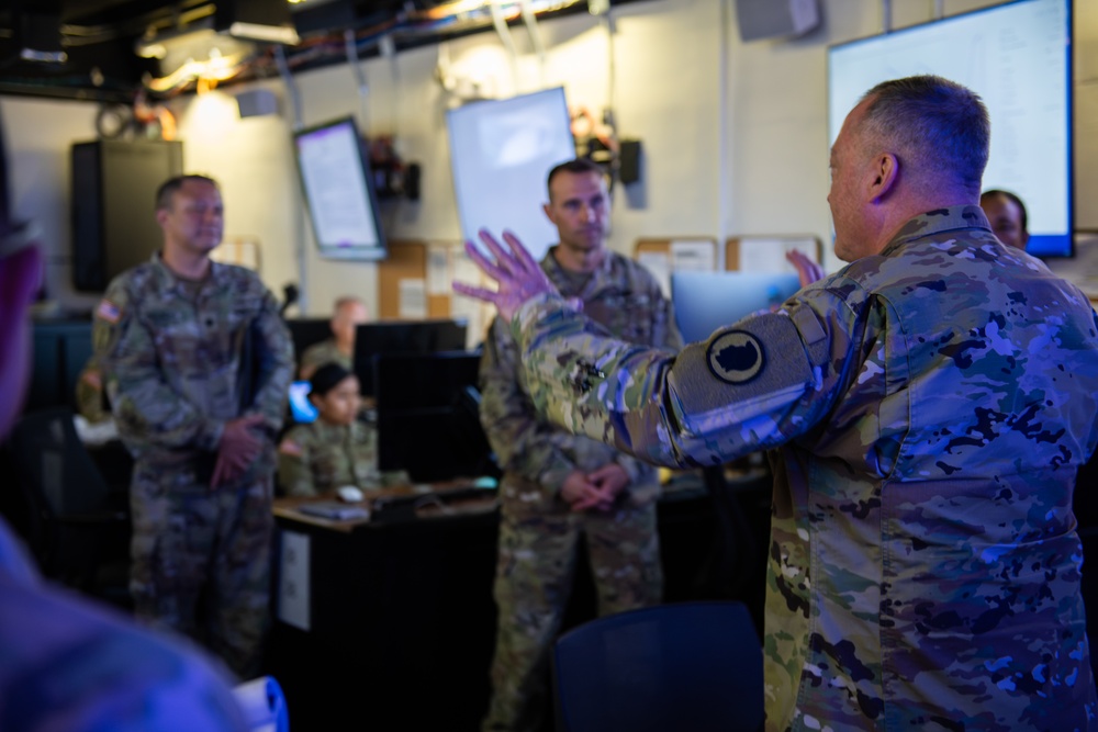 A group of U.S. Army soldiers in MultiCam/OCP camouflage uniforms are gathered in what appears to be a military operations center or command post. A senior soldier in the foreground is gesturing with both hands, apparently briefing or instructing the others. The room contains multiple computer workstations, monitors displaying information, and wall-mounted screens.