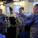 A group of U.S. Army soldiers in MultiCam/OCP camouflage uniforms are gathered in what appears to be a military operations center or command post. A senior soldier in the foreground is gesturing with both hands, apparently briefing or instructing the others. The room contains multiple computer workstations, monitors displaying information, and wall-mounted screens.
