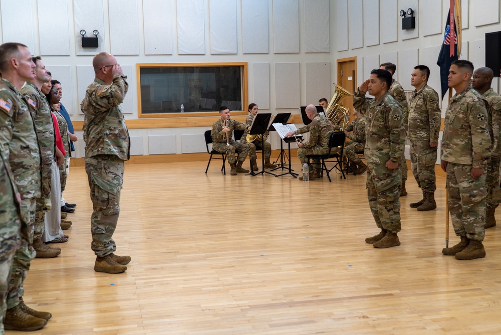 A U.S. Army military ceremony is taking place indoors in what appears to be a formal hall or gymnasium with hardwood floors. Soldiers in MultiCam/OCP camouflage uniforms are arranged in formation on both sides while a small brass ensemble performs in the background. At least two soldiers are rendering salutes, suggesting a formal event such as a change of command, promotion, or award ceremony.