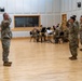 A U.S. Army military ceremony is taking place indoors in what appears to be a formal hall or gymnasium with hardwood floors. Soldiers in MultiCam/OCP camouflage uniforms are arranged in formation on both sides while a small brass ensemble performs in the background. At least two soldiers are rendering salutes, suggesting a formal event such as a change of command, promotion, or award ceremony.