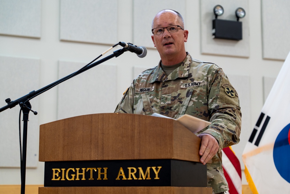 A U.S. Army soldier with the name tape 'ROGERS' is speaking at a podium labeled 'Eighth Army' during an official military ceremony or event. He is wearing MultiCam (OCP) combat uniform with U.S. Army insignia and what appears to be an Eighth Army patch on his right shoulder. The South Korean and American flags are visible in the background, indicating this event likely takes place in South Korea.