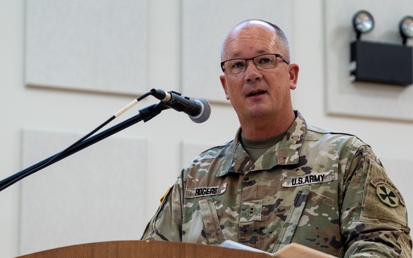 A U.S. Army soldier with the name tape 'ROGERS' is speaking at a podium labeled 'Eighth Army' during an official military ceremony or event. He is wearing MultiCam (OCP) combat uniform with U.S. Army insignia and what appears to be an Eighth Army patch on his right shoulder. The South Korean and American flags are visible in the background, indicating this event likely takes place in South Korea.