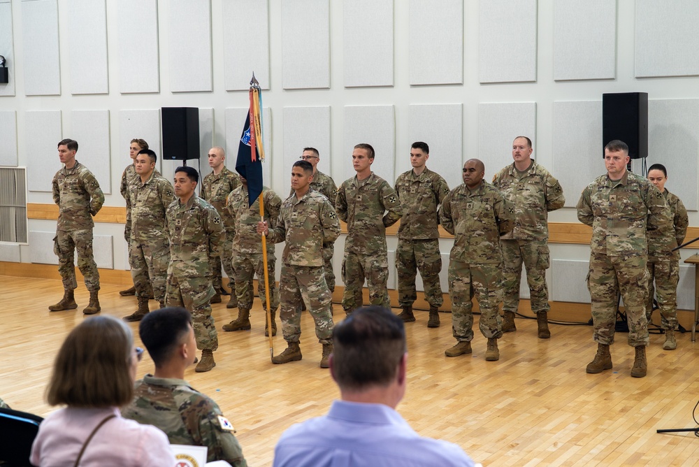 A group of approximately 13 military personnel in U.S. Army OCP (Operational Camouflage Pattern) uniforms are standing in formation on a wooden gymnasium or auditorium floor. One soldier in the center is holding a unit guidon (flag on a staff with blue, red, and gold colors). An audience is seated in the foreground observing what appears to be a formal military ceremony.