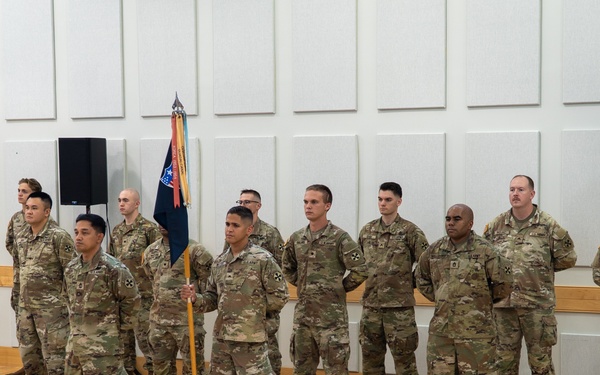 A group of approximately 13 military personnel in U.S. Army OCP (Operational Camouflage Pattern) uniforms are standing in formation on a wooden gymnasium or auditorium floor. One soldier in the center is holding a unit guidon (flag on a staff with blue, red, and gold colors). An audience is seated in the foreground observing what appears to be a formal military ceremony.