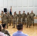 A group of approximately 13 military personnel in U.S. Army OCP (Operational Camouflage Pattern) uniforms are standing in formation on a wooden gymnasium or auditorium floor. One soldier in the center is holding a unit guidon (flag on a staff with blue, red, and gold colors). An audience is seated in the foreground observing what appears to be a formal military ceremony.