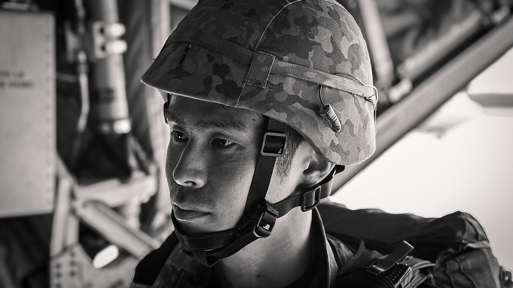 A black-and-white portrait photograph of a young male soldier wearing a camouflage-patterned helmet with chin strap secured. He appears to be seated inside a military vehicle or aircraft, with structural metal framework visible in the background. His expression is serious and focused, suggesting an operational or training context.