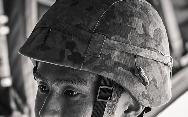 A black-and-white portrait photograph of a young male soldier wearing a camouflage-patterned helmet with chin strap secured. He appears to be seated inside a military vehicle or aircraft, with structural metal framework visible in the background. His expression is serious and focused, suggesting an operational or training context.
