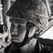 A black-and-white portrait photograph of a young male soldier wearing a camouflage-patterned helmet with chin strap secured. He appears to be seated inside a military vehicle or aircraft, with structural metal framework visible in the background. His expression is serious and focused, suggesting an operational or training context.